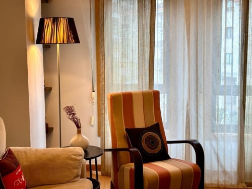 Luminoso salón del apartamento Baiona con silla naranja a rayas, lámpara de pie y vistas de la ciudad a través de cortinas translúcidas