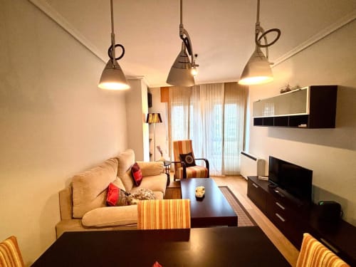Moderno y luminoso salón de apartamento en Baiona con sofá beige, lámparas colgantes, TV de pantalla plana y zona de comedor.