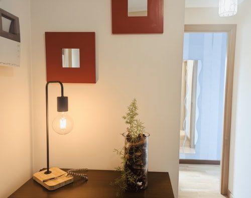 Entrada moderna del apartamento Holibai Baiona con lámpara minimalista negra, planta en maceta y paneles de arte rojo en la pared