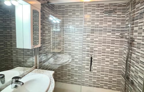 Baño moderno en apartamento Holibai, Baiona, con paredes de azulejos a rayas, ducha de cristal y lavabo blanco con espejo