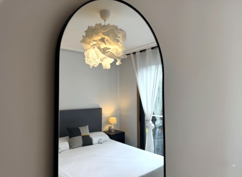 Espejo moderno del dormitorio reflejando cama con ropa de cama blanca, cabecera oscura, lámpara colgante y ventana con cortinas en Holibai