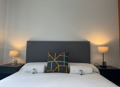 Dormitorio moderno en apartamento Holibai de Baiona con cabecero oscuro, ropa de cama blanca, almohada geométrica e iluminación cálida