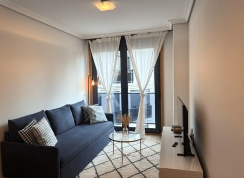 Moderno salón con sofá azul, mesa de cristal y balcón con vistas a la playa de Baiona en apartamento Holibai