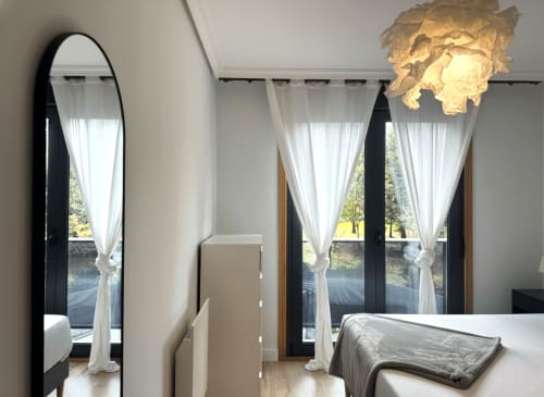 Dormitorio moderno en apartamento Holibai Cousiña de Baiona con espejo alto, lámpara colgante elegante y ventanales de piso a techo