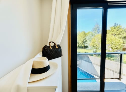 Elegante dormitorio con vistas a la piscina en Holibai, Baiona. Sombrero de paja y bolso negro en estante, puertas francesas con vistas a la piscina y