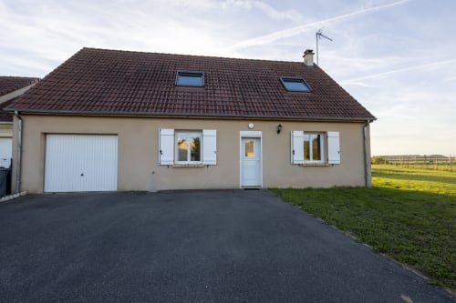 Charmant cottage Accolade à Châteaudun avec vue sur le jardin, façade beige, volets blancs, toiture en tuiles sombres, garage et allée pavée.