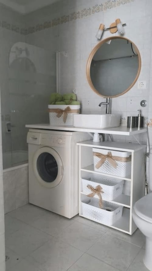 Baño moderno con espejo redondo de madera, lavabo blanco y lavadora en estudio frente al mar de MiramaRent