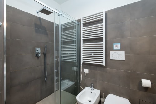 Modernes Badezimmer im Magnolia Apartments Mailand mit grauen Fliesen, Glasduschabtrennung, weißem Waschbecken, Bidet und Handtuchheizkörper
