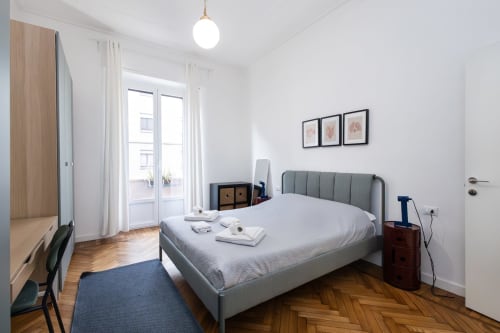 Dormitorio moderno en Magnolia Apartments Milan con cama tapizada gris, suelo de parqué y balcón con vista al jardín