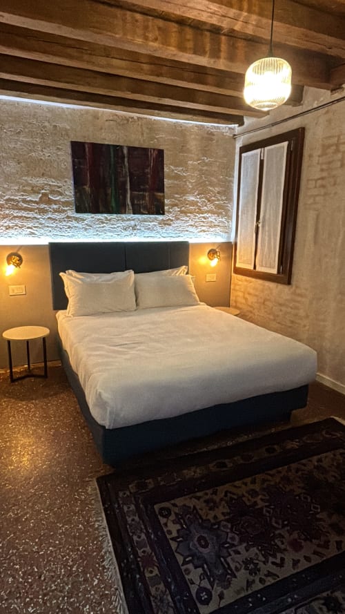 Accogliente letto matrimoniale con biancheria bianca nella camera dell'hotel Casanova Hideaway Venice, con travi a vista e illuminazione calda