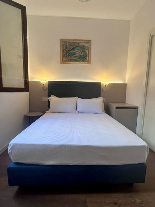 Letto matrimoniale moderno nella camera dell'hotel Casanova Hideaway Venice con biancheria bianca, testata scura, applique murali e quadri incorniciati