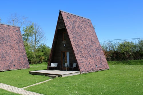 Modern A-frame lodge with red brick exterior and wooden deck at Domaine des Piéris in Grandcamp-Maisy, surrounded by green