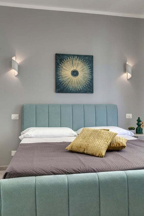 Modern bedroom at Palazzo Belvedere with sage green headboard, yellow pillows, sunburst artwork, and ambient wall lighting.
