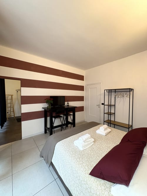 Camera moderna dell'hotel Le Arche a Verona con parete a righe, scrivania, letto con biancheria bianca e appendiabiti nero
