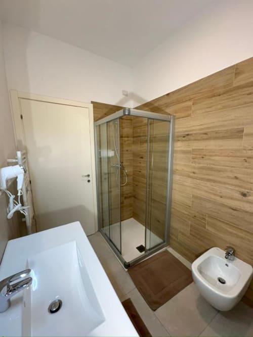 Bagno moderno dell'appartamento Le Arche Verona con doccia in vetro, lavabo bianco, bidet e rivestimento murale effetto legno