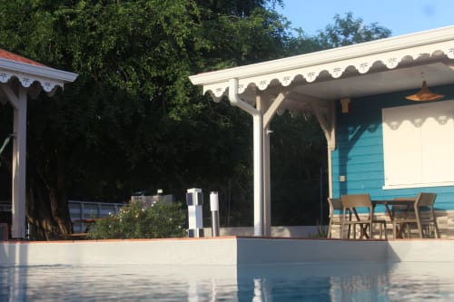 Vue de la terrasse couverte et de la piscine d'une villa bleue au style créole, entourée de végétation.