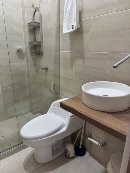 Baño moderno del hotel Olamar Living en Santiago de Tolú con inodoro, lavabo redondo, ducha y paredes de azulejos color beige