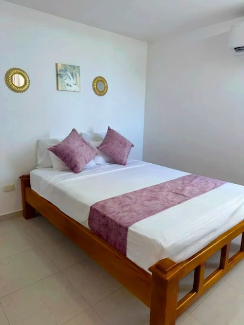 Habitación limpia del hotel Olamar Living en Santiago de Tolú con cama de madera, ropa de cama blanca, almohadas y camino burgundy, y pared