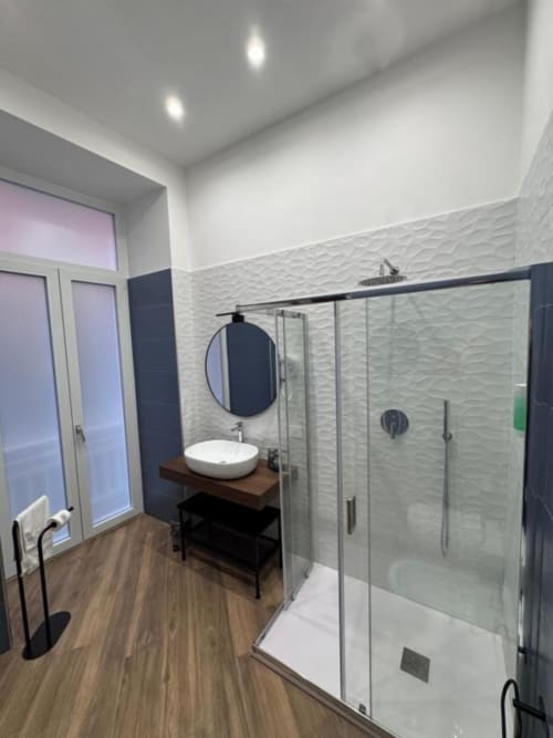 Bagno moderno con piastrelle bianche texturizzate, specchio rotondo nero, lavabo da appoggio su mobile in legno e doccia con box in vetro