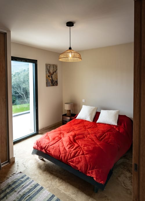 Chambre moderne à La Conciergerie du Cap Corse avec lit matelassé rouge, suspension en rotin et vue sur les montagnes