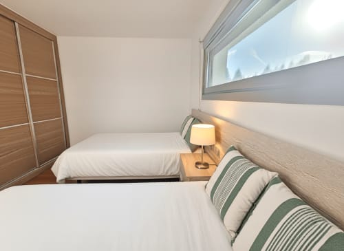 Moderno dormitorio en apartamento de Baiona con camas gemelas, ventana con vistas al mar, armario de madera y lámpara de lectura