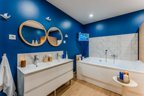 Modern blue bathroom at Wellkhome Bordeaux with marble tiles, double sink vanity, soaking tub, and round wooden mirrors