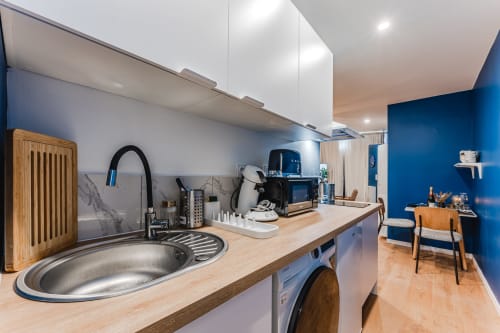 Modern kitchenette at Wellkhome Bordeaux with blue accent wall, stainless steel sink, coffee maker, and wooden countertop
