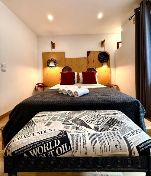 Quarto aconchegante da pousada Kindnessmoon em Óbidos com colcha estampada em jornal, almofadas vermelhas e cabeceira de madeira