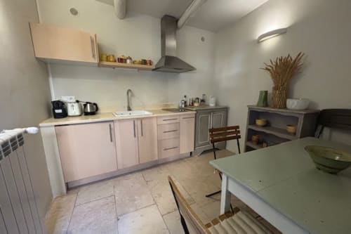 Modern kitchen in Lourmarin courtyard apartment with light cabinetry, stainless steel hood, dining area, and tile flooring.