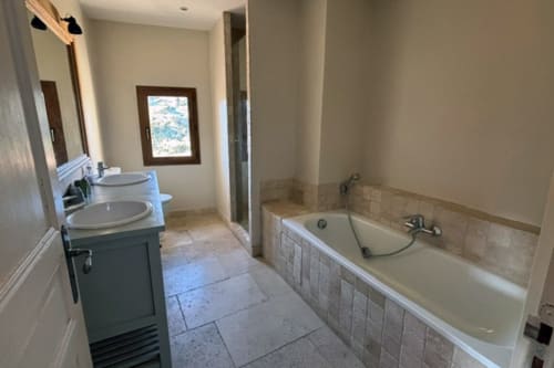 Spacious bathroom in Lourmarin apartment with dual sinks, bathtub with tile surround, and window view