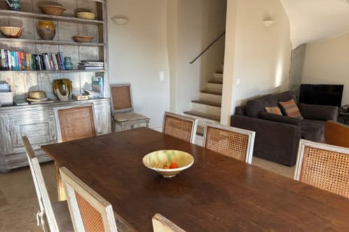 Dining area in Lourmarin apartment with dark wood table, rattan chairs, built-in shelving, and staircase access to upper