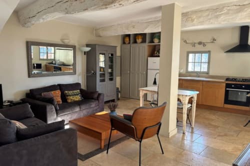 Open-plan living area in Lourmarin apartment with black sofas, wooden table, kitchenette, exposed beams, and countryside