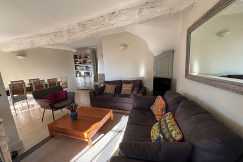 Spacious living room in Lourmarin apartment with dark sofa, wooden beams, mirror, dining area, and exposed stone ceiling
