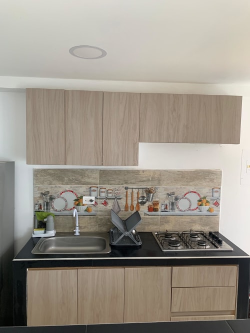 Kitchenette moderno con encimera negra, estufa de gas, fregadero y muebles de madera clara en apartamento Olamar Living en Coveñas