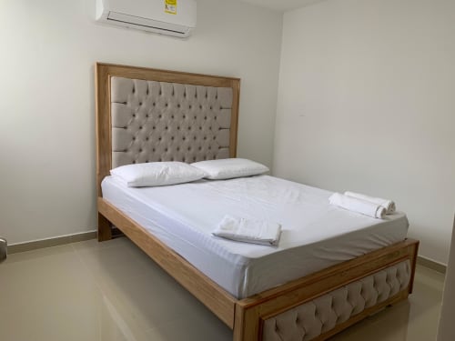 Cama de madera con cabecero y pie tapizado en beige, ropa de cama blanca, almohadas y toallas enrolladas en una habitación de hotel impecable