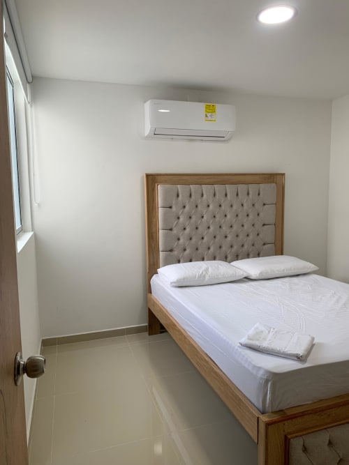 Dormitorio moderno en Olamar Living, Coveñas: cama de madera, ropa de cama blanca, aire acondicionado y piso de cerámica