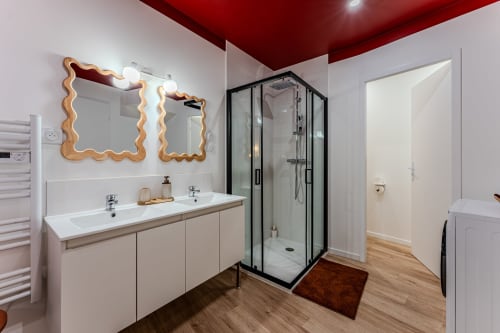 Modern bathroom at Wellkhome Bordeaux with red ceiling, wavy wooden mirrors, double vanity, and black-framed shower enclosure