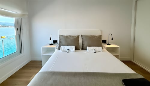 Dormitorio moderno con vistas al mar en Baiona, España. Cama blanca con almohadas de oliva, mesitas de noche, suelos de madera y ventana