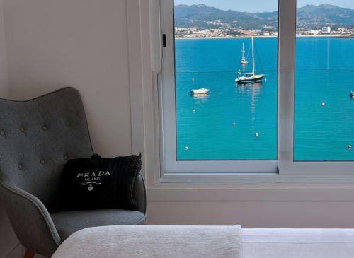 Sillón gris con bolso Prada junto a ventana con vista a bahía turquesa con veleros, costa de Baiona y montañas