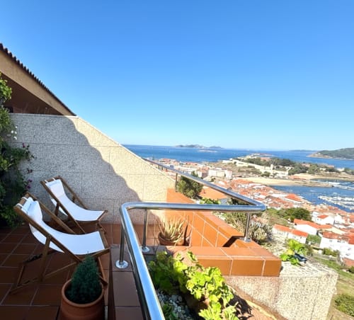 Terraza del apartamento en Baiona con tumbonas vista al mar, pueblo costero y océano azul abajo, plantas en macetas y barandilla