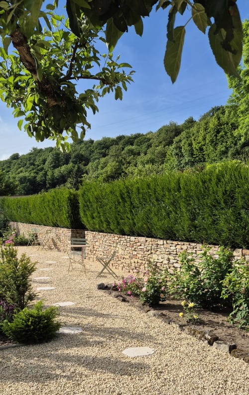 Grindtuin-patio bij Moulin Renaudiots met stenen muur, bloeiende planten en weelderige groene helling in Autun, Frankrijk