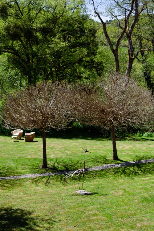 Weelderige tuin bij Moulin Renaudiots in Autun met verzorgde gazon, metalen sculptuur en bloeiende struiken onder hoge bomen.