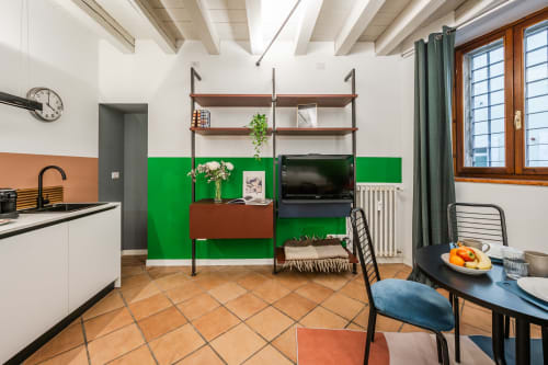 Zona soggiorno di moderno appartamento a Verona con parete verde accento, angolo cottura, mensole, TV, travi in legno e tavolo da pranzo