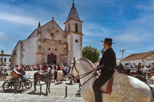 Homem em cavalo branco na festa equestre de Golegã com igreja tradicional e carroças de cavalos na praça