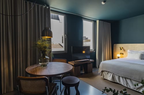 Estudio moderno de hotel en Oporto con mesa redonda, cama, TV, iluminación colgante y vistas de la ciudad a través de grandes ventanales