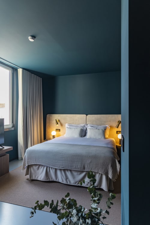 Estudio de hotel moderno en Oporto con paredes azul petróleo, cabecero tapizado beige, ropa de cama blanca e iluminación cálida