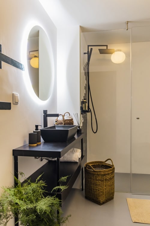 Baño moderno en estudio de hotel en Oporto con tocador negro, espejo ovalado iluminado, ducha de cristal, plantas y cesto de almacenamiento tejido