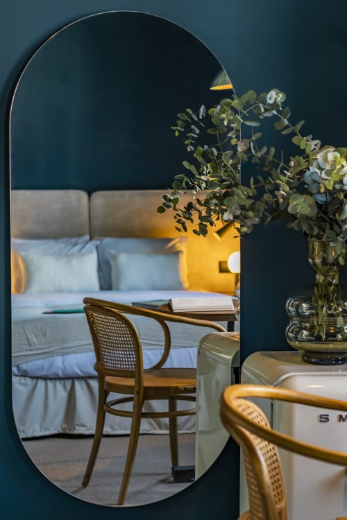Espejo de hotel reflejando dormitorio con silla de madera, flores de eucalipto y lámpara amarilla en estudio compacto de Oporto