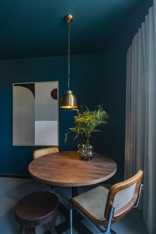 Comedor moderno en estudio de hotel de Oporto con mesa redonda de nogal, sillas de ratán, paredes verde azulado, lámpara colgante dorada y decoración abstracta