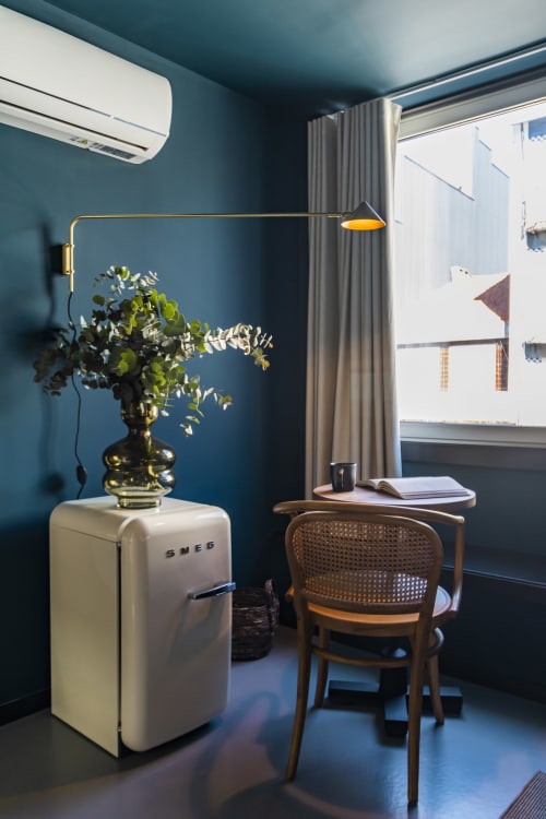 Estudio compacto moderno en Oporto con paredes verde azulado, refrigerador SMEG, silla de madera, lámpara de escritorio de latón y vista de la ciudad